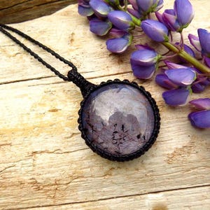 May include: A round, dark purple gemstone pendant encased in a black macrame cord setting. The pendant hangs from a matching black braided cord necklace, featuring a smooth, polished surface with visible internal inclusions and dark veining.