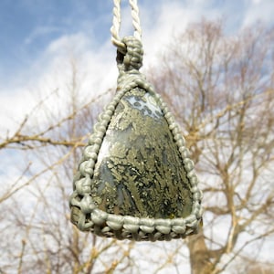 Puede incluir: Un colgante triangular con una piedra gris y dorada engastada en un nudo macramé. La piedra tiene un patrón único de motas doradas.