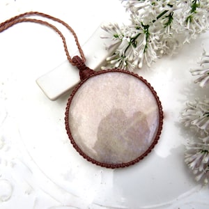 May include: A round, iridescent, light-colored stone pendant encased in a brown macrame cord setting. The pendant hangs from a matching brown, twisted cord necklace. The stone has a smooth, polished surface with subtle pearlescent reflections.