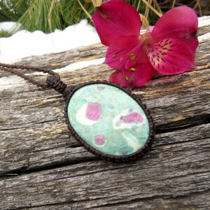 Può includere: Un ciondolo ovale verde e rosa con una collana in corda marrone. Il ciondolo è fatto di una pietra naturale con un motivo unico. La collana è appoggiata su un pezzo di legno invecchiato.