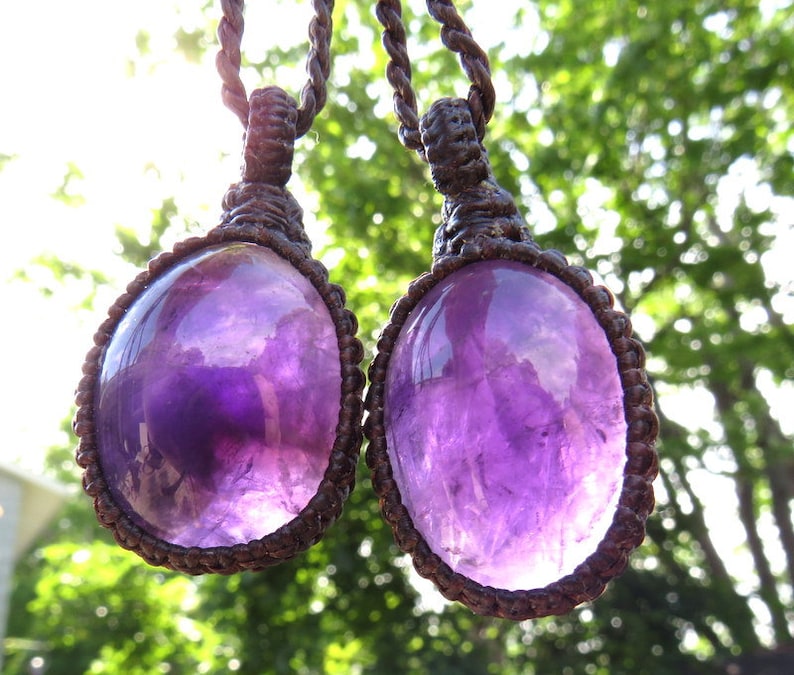 May include: Two amethyst gemstone pendants on brown cord necklaces. The gemstones are oval shaped and have a purple hue. The necklaces are knotted and have a macrame style.