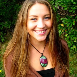 May include: An oval-shaped pendant featuring a vibrant magenta and green stone set in a black woven cord necklace. The pendant displays a smooth, polished surface with distinct colour variations, suspended from a simple, dark, adjustable-style cord.
