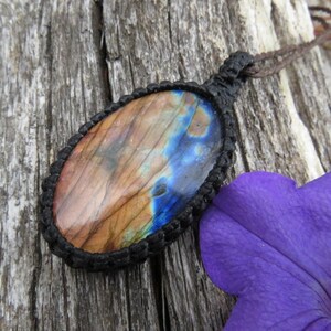 May include: A brown and blue labradorite gemstone pendant with a black cord. The pendant is oval shaped and is set in a black macrame knot.
