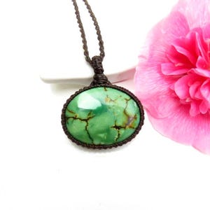Könnte beinhalten: Eine grüne ovale Edelstein-Anhänger-Halskette mit einer braunen geflochtenen Kordel. Der Edelstein hat braune Adern und ist in einen braunen geflochtenen Rahmen gefasst. Eine rosa Blume befindet sich im Hintergrund.