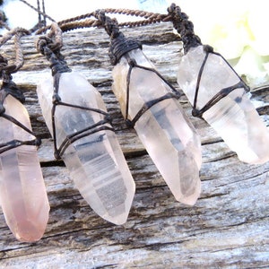 Puede incluir: Cinco puntas de cristal de cuarzo transparente envueltas en cordón marrón, colgando de una superficie de madera. Los cristales son de varios tamaños y formas, algunos muestran un ligero tono rosado.