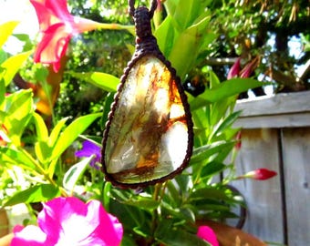 May include: A clear quartz crystal pendant with brown inclusions, wrapped in brown cord, hanging from a branch in a garden setting.