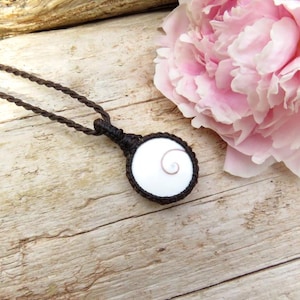 Peut inclure: Un collier artisanal avec un pendentif blanc orné d'un motif en spirale, serti dans un cadre tissé marron foncé. Le collier est suspendu à un cordon marron assorti. Le fond comprend une surface en bois et une pivoine rose.