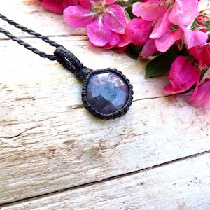 May include: A necklace featuring a round, faceted dark purple gemstone set in a black macrame cord frame. The pendant is attached to a twisted black cord necklace, showcasing a detailed knotted design around the stone's edge and a secure loop.