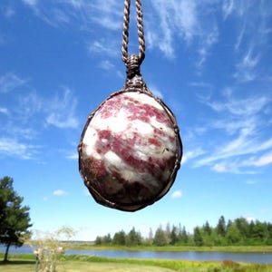 May include: A pendant necklace featuring a large, round gemstone with a mix of white and deep red hues. The stone is encased in a metal wire cage and suspended from a brown cord. The background shows a blue sky with clouds and a landscape.
