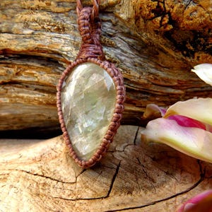 May include: A teardrop-shaped pendant featuring a translucent, pale green gemstone with internal inclusions. The stone is encased in a brown macrame cord setting with a woven loop at the top, showcasing intricate knotting and a textured finish.