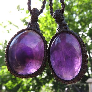 May include: Two amethyst gemstone pendants on brown cord necklaces. The amethyst stones are oval shaped and have a deep purple color.