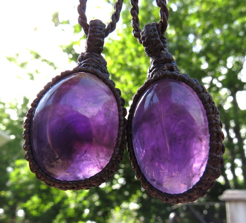 May include: Two amethyst gemstone pendants on brown cord necklaces. The amethyst stones are oval shaped and have a deep purple color.