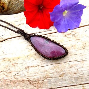 Puede incluir: Un collar artesanal con un colgante de piedra preciosa púrpura pulida, de forma ovalada. El colgante está engastado en un marco de macramé marrón y suspendido de un cordón a juego. Flores rojas y moradas descansan cerca del collar sobre una superficie de madera clara.