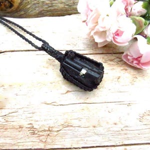 Puede incluir: Un collar con colgante negro con un colgante rectangular texturizado. El colgante tiene un pequeño acento cuadrado de color plateado. El collar está sobre una superficie de madera clara, con flores rosas en el fondo.