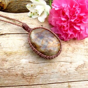 May include: A handmade macramé necklace with a large, oval stone pendant. The stone displays brown, grey, and gold hues. The pendant is set in a woven, brown macramé casing and is suspended from a matching cord.