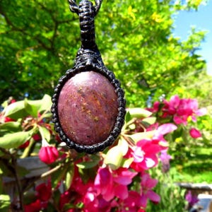 May include: An oval-shaped pendant featuring a polished, deep reddish-purple stone with subtle golden inclusions. The stone is encased in a black macrame cord setting with a braided loop at the top, designed to be worn as a necklace pendant.