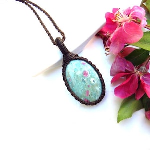 May include: An oval-shaped pendant featuring a mottled light green and pink stone set in a dark brown knotted cord frame. The pendant hangs from a matching dark brown braided cord necklace, showcasing a natural stone with distinct pink inclusions.