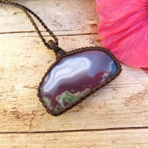 Puede incluir: Un collar de cordón de cuero marrón con un colgante de ágata púrpura y blanco. El colgante está envuelto en cordón marrón y tiene un aspecto natural y orgánico.