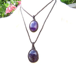 May include: Two amethyst gemstone pendants on brown cord necklaces. The larger pendant is at the bottom of the necklace and the smaller pendant is at the top.