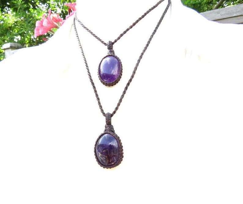 May include: Two amethyst gemstone pendants on brown cord necklaces. The larger pendant is at the bottom of the necklace and the smaller pendant is at the top.