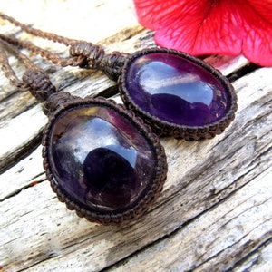 May include: Two amethyst gemstone pendants on brown cord necklaces. The gemstones are oval shaped and have a deep purple color with a slight shimmer.