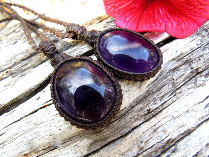 May include: Two amethyst gemstone pendants on brown cord necklaces. The gemstones are oval shaped and have a deep purple color with a slight shimmer.