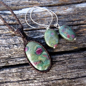 May include: A green and pink ruby zoisite necklace and earrings set. The necklace features a large oval-shaped stone with a brown cord. The earrings are silver hoops with oval-shaped stones.