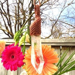 Puede incluir: Un colgante de cristal de cuarzo transparente con un envoltorio de cordón de cuero marrón. El cristal es transparente con inclusiones de otros minerales, y el cordón está envuelto en un nudo decorativo. El colgante cuelga de un ramo de flores.
