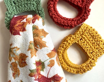 May include: Three crochet towel holders in green, red, and yellow. One green holder is attached to a white kitchen towel featuring a repeating pattern of orange, red, and brown autumn leaves. The holders are circular with a rectangular top tab.