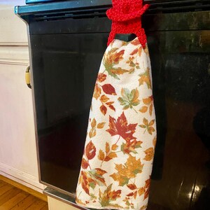 May include: Red crocheted oven mitt with a leaf patterned kitchen towel hanging on an oven door handle.