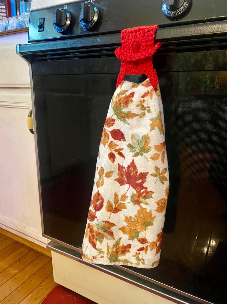 May include: Red crocheted oven mitt with a leaf patterned kitchen towel hanging on an oven door handle.