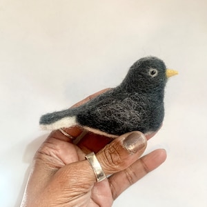May include: A needle-felted bird figurine crafted from dark charcoal gray wool fibers. The bird features a small, pointed yellow beak, a round black eye, and a distinct white patch of wool along the underside of its tail feathers.