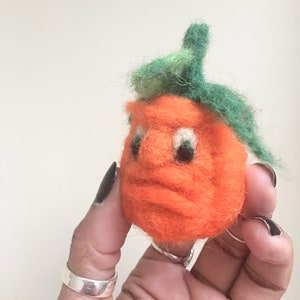 May include: A handmade orange felt pumpkin with a face and green felt leaves. The pumpkin has black eyes and a nose. The pumpkin is held in a hand with silver rings and black nail polish.