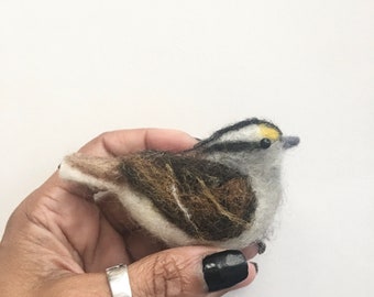 Könnte beinhalten: Handgefertigte Vogelfigur aus Filz. Der Vogel besteht aus einer Mischung aus brauner, weißer, grauer und gelber Wolle. Der Körper des Vogels ist eine Mischung aus Braun und Weiß, mit einem grauen Kopf und einem gelben Fleck oben. Der Vogel sitzt auf einer weißen Oberfläche.