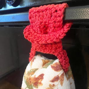 May include: A red crocheted towel holder with a floral patterned kitchen towel hanging from it. The towel holder is attached to a black oven door.
