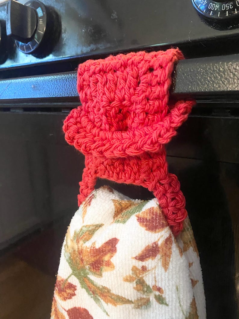 May include: A red crocheted towel holder with a floral patterned kitchen towel hanging from it. The towel holder is attached to a black oven door.