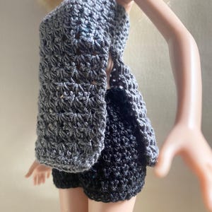 May include: A doll wearing a handmade crochet outfit. The outfit includes a gray halter top and a matching open-front jacket, paired with black shorts. The crochet work has a textured appearance.