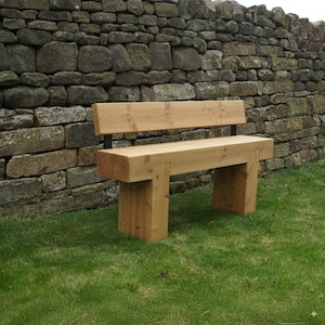 May include: A light-brown wooden bench with a backrest, constructed from thick planks. The bench is supported by square legs. The background features a stone wall and green grass.