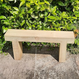 May include: A simple wooden bench with a natural finish. The bench is made of solid wood and has a rectangular shape. It is sitting on a concrete surface.