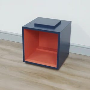 May include: A navy blue cube-shaped side table with a coral interior. The table has a small, raised rectangular top. The table is made of wood and sits on a light-coloured wood floor.