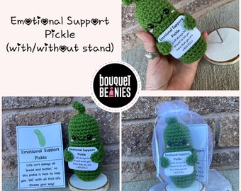 May include: A green crocheted pickle with a smiling face, holding a card that says "Emotional Support Pickle" and "I will always be around to let you know that you are a big dill!" The pickle is sitting on a wooden stand.