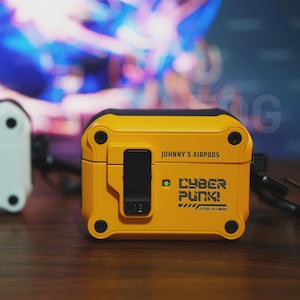 Può includere: Custodia gialla e nera per AirPods con la scritta "JOHNNY'S AIRPODS" e "CYBER PUNK! FUTURE IS COMING" stampata sul davanti. La custodia ha una forma rettangolare con angoli arrotondati e un fermaglio nero.