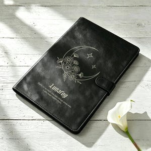 Pode incluir: Um caderno retangular preto com fecho magnético. A capa apresenta um desenho de linhas brancas de uma lua crescente preenchida com margaridas e estrelas. Abaixo do design, o texto diz: "Lunafog. Don't try so hard; the best things come when you least expect them."