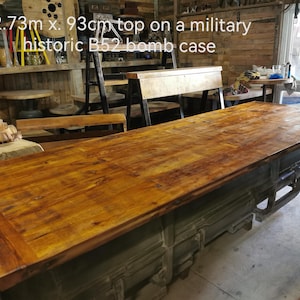 May include: A large, rectangular wooden table with a distressed finish, measuring approximately 2.73 metres long and 93 centimetres wide. The table rests on a vintage military bomb case, painted in a muted green colour. The text on the image reads: 2.73m x 93cm top on a military historic B52 bomb case.