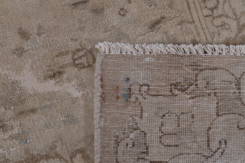 May include: Close-up of a beige rug with a fringe border. The rug has a subtle pattern of faded floral designs.