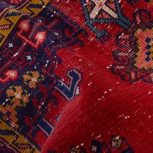May include: Close-up of a red, patterned rug with a detailed design. The rug features a border with yellow and navy blue geometric patterns. The central area is predominantly red with intricate floral and geometric motifs in various colors.
