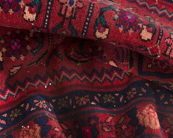 May include: Close-up of a richly detailed, folded rug. The rug features a deep red background with intricate patterns in navy blue, green, and touches of gold. The design includes floral and geometric motifs, creating a textured and ornate appearance.