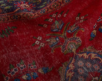 May include: Close-up of a distressed red rug with intricate floral patterns in blue, green, and white. The rug has a faded, vintage appearance, with visible wear and tear, suggesting age and use. The design includes detailed borders and central motifs.