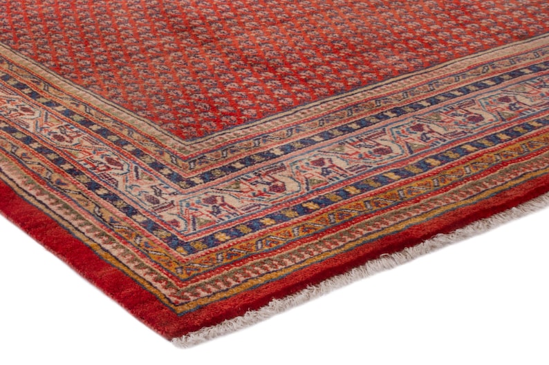 Puede incluir: Esquina de una alfombra oriental roja con un patr&oacute;n floral detallado. La alfombra tiene un borde con dise&ntilde;os intrincados en azul, beige y dorado. El borde de la alfombra est&aacute; terminado con flecos.