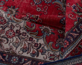 May include: Close-up of a vintage red rug with intricate floral patterns in cream, blue, and brown. The rug has a distressed look, with visible wear and tear, and a dark blue border. The rug is folded over, revealing its layered construction.